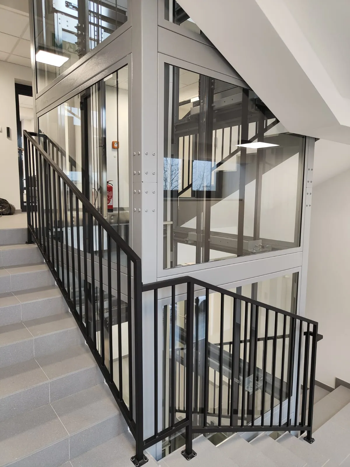 Jasnoszary przeszklony szyb windy w klatce schodowej z czarnymi balustradami