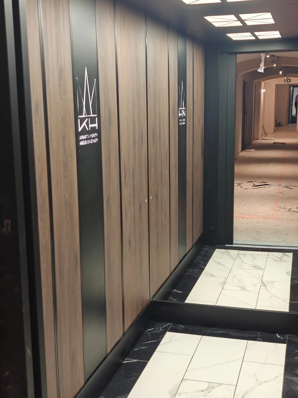 Kabina z drewnopodobnymi panelami i podświetlanym logo Apartamentów Królewskie Huty, otwarte drzwi na korytarz